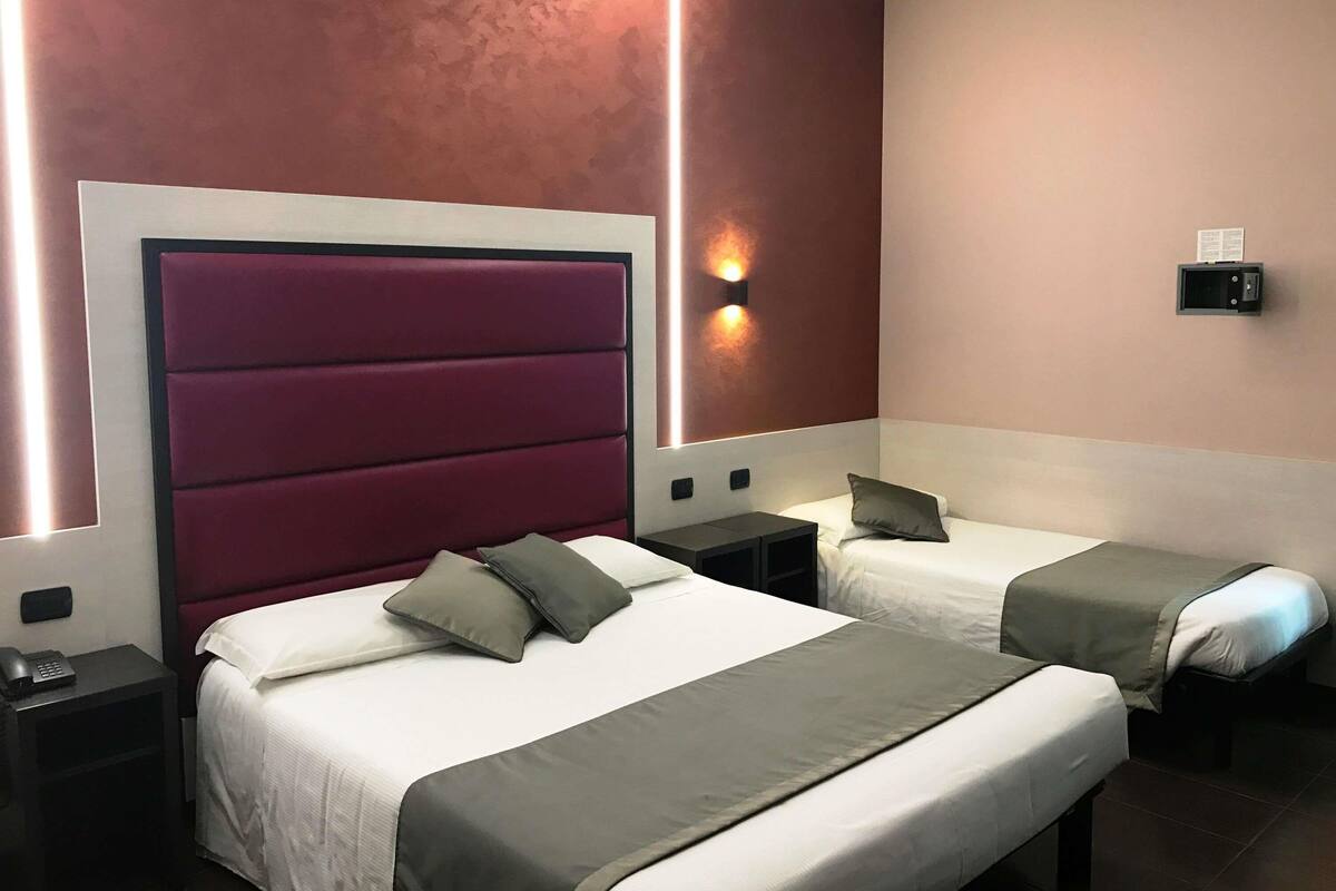 A hotel room with two beds, a large padded headboard, bedside tables, and wall-mounted lighting.