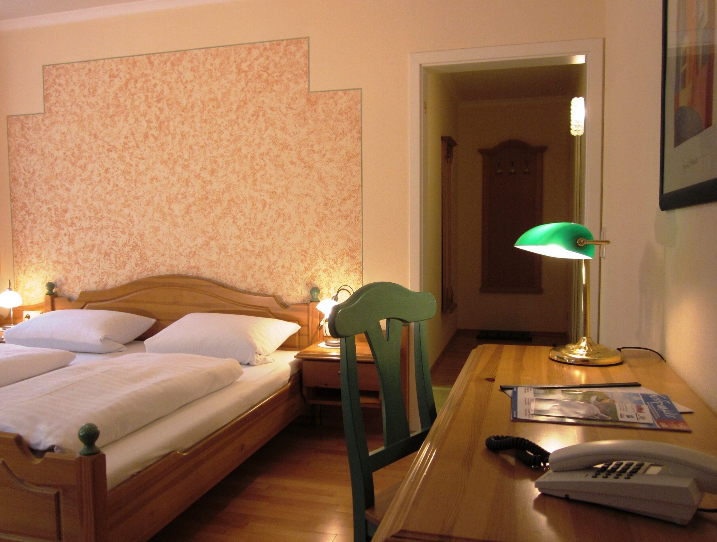 A hotel room with a wooden bed, a green chair, a desk with a lamp, and a telephone.