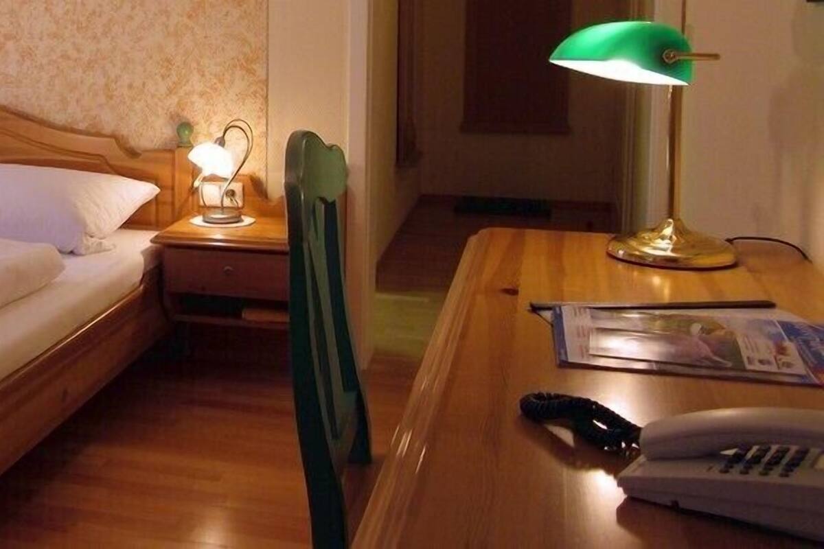 Una habitación de hotel con una cama de madera, una mesita de noche con lámpara, una silla y un escritorio con teléfono y control remoto.