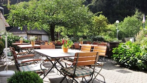Un patio al aire libre con mesas y sillas de madera, rodeado de follaje y un edificio al fondo.