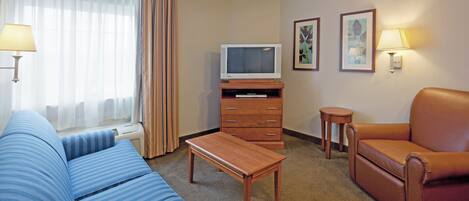 A hotel room with a blue sofa, a brown leather chair, a wooden coffee table, a TV on a wooden stand, and two wall-mounted lamps.