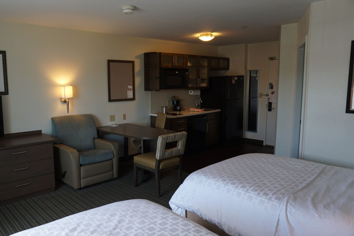 A hotel room with two beds, a desk with a chair, a television, a kitchenette, and a framed picture on the wall.