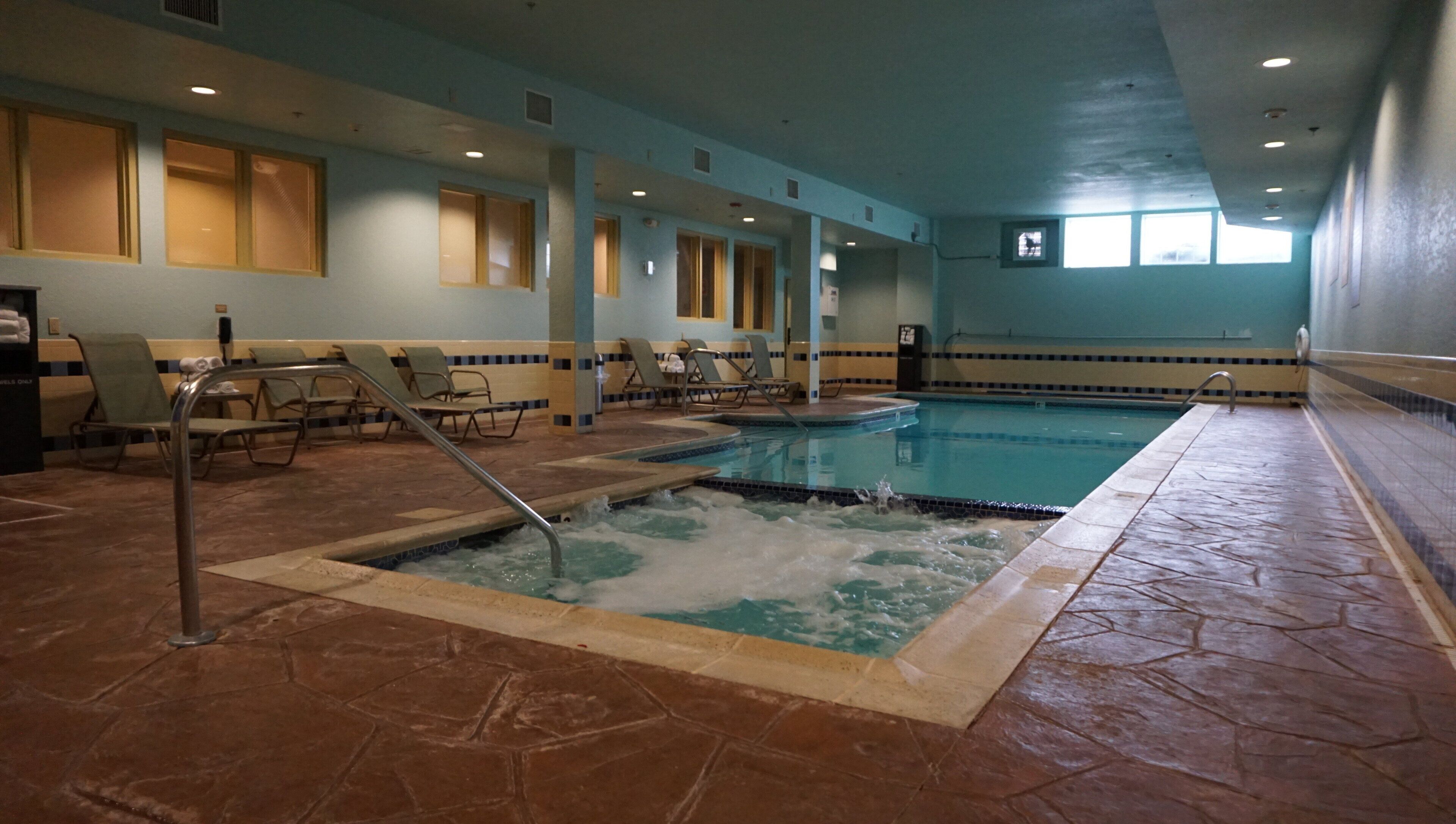 An indoor swimming pool with a hot tub, lounge chairs, and a tiled floor.