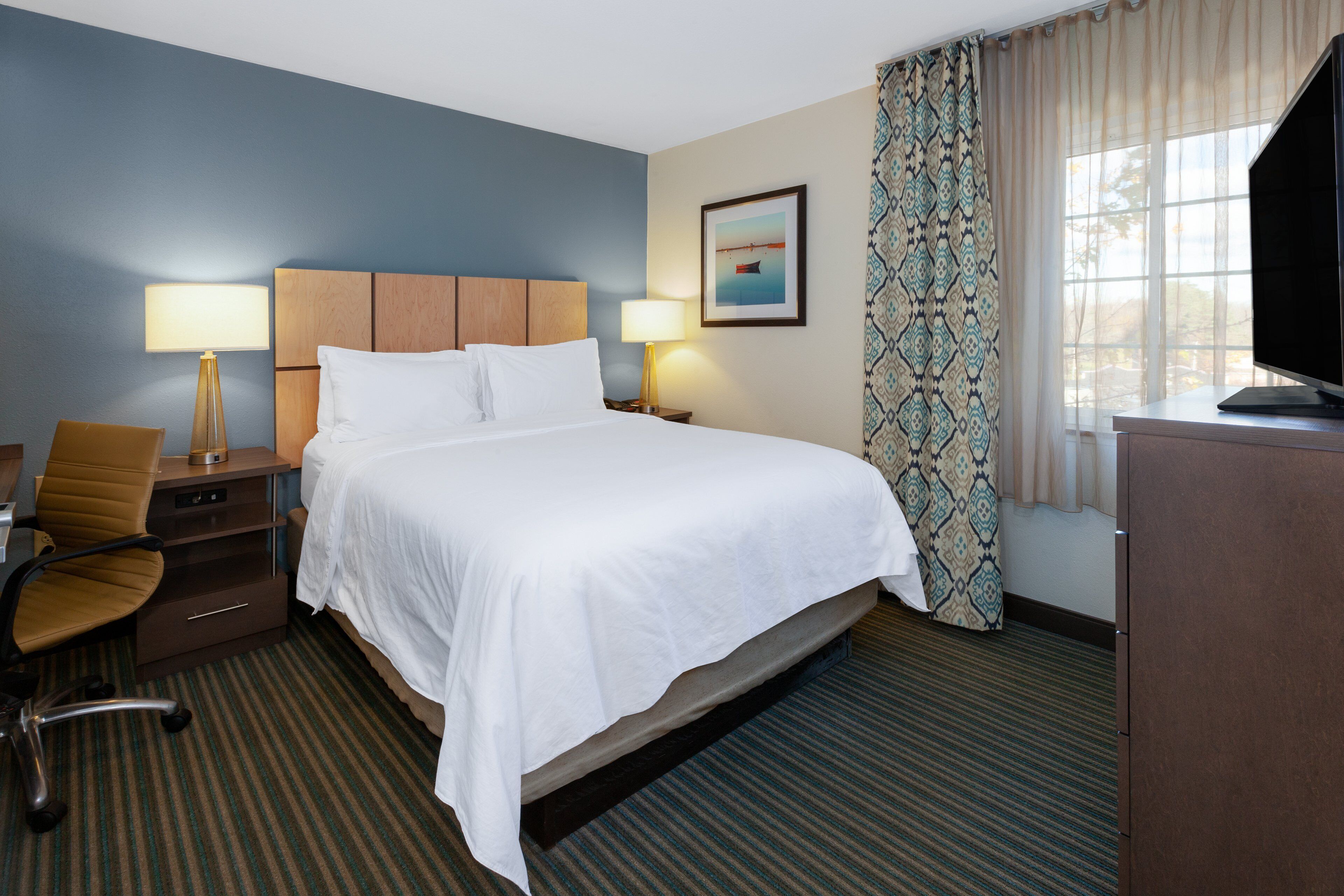 A hotel room with a large bed, two bedside lamps, a desk with a chair, a television, and a window with patterned curtains.