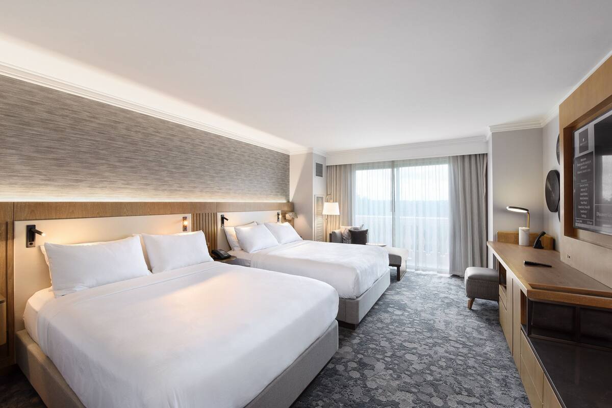 A hotel room with two beds, a desk, a chair, a TV, and a large window with curtains.