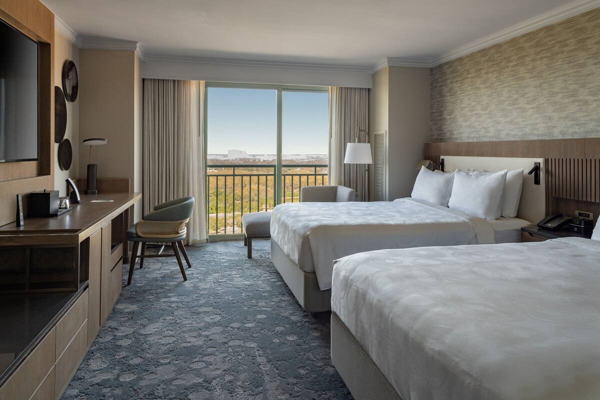 A hotel room with two beds, a desk, a chair, a TV, and a balcony with a view.