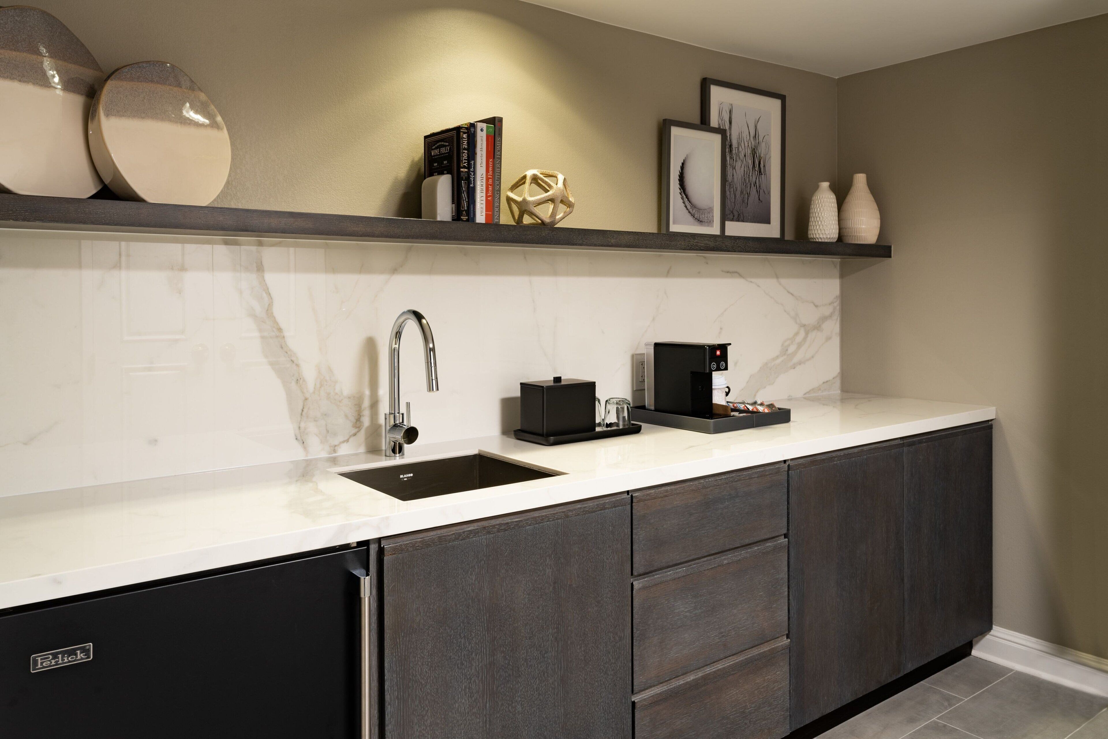 Een moderne keuken met een geïntegreerde vaatwasser, donkere kastjes, een witte marmeren aanrecht en een plank met decoratieve objecten.