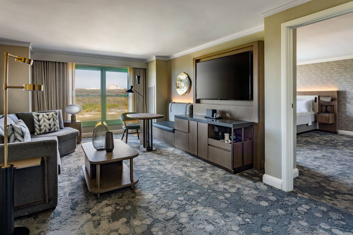 A hotel room with a sofa, a small table, a TV, and a view of the outdoors.