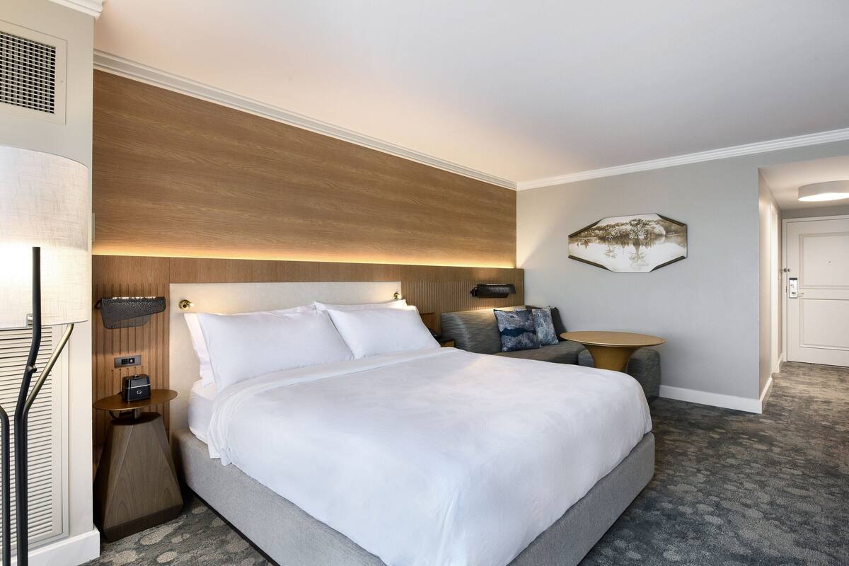 A hotel room with a large bed, a bedside table with a lamp, a small round table, and a wall-mounted artwork.