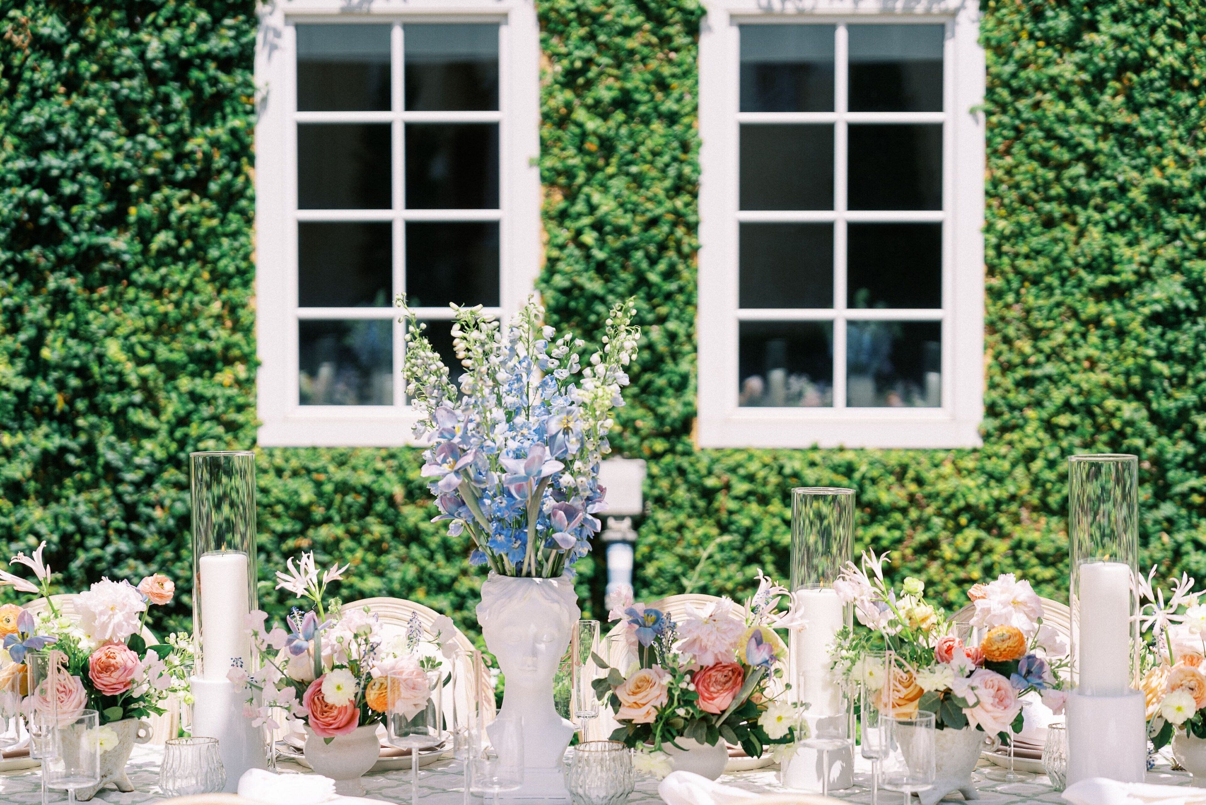 A table set with floral arrangements and candles, set against a backdrop of a building with large windows and a green ivy-covered wall.