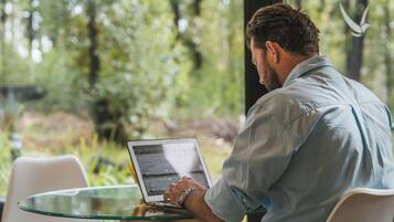 Una persona sentada a una mesa de vidrio con una computadora portátil, al aire libre con follaje de fondo.