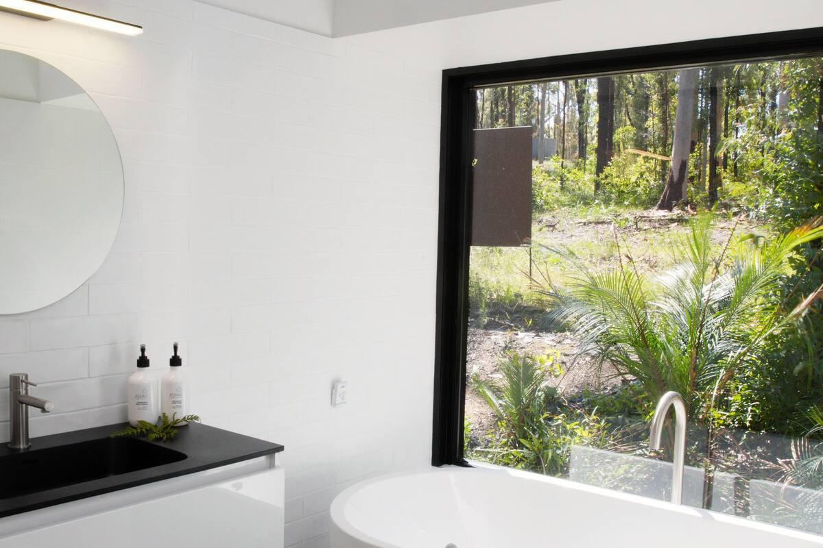 A modern bathroom with a freestanding bathtub, a black countertop, a round mirror, and a view of a forest through a large window.