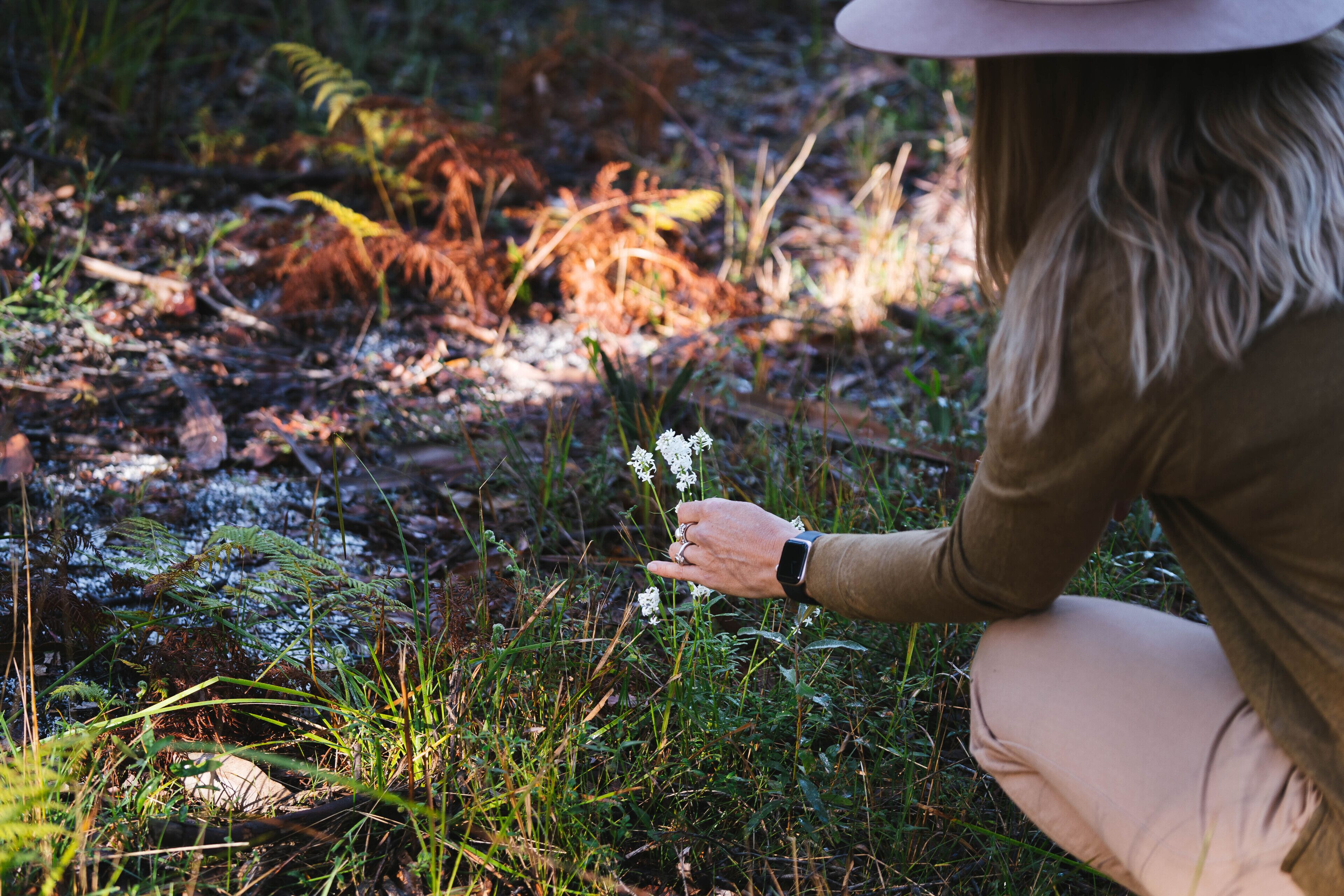 A person crouching in a natural setting with greenery and a small white flower.