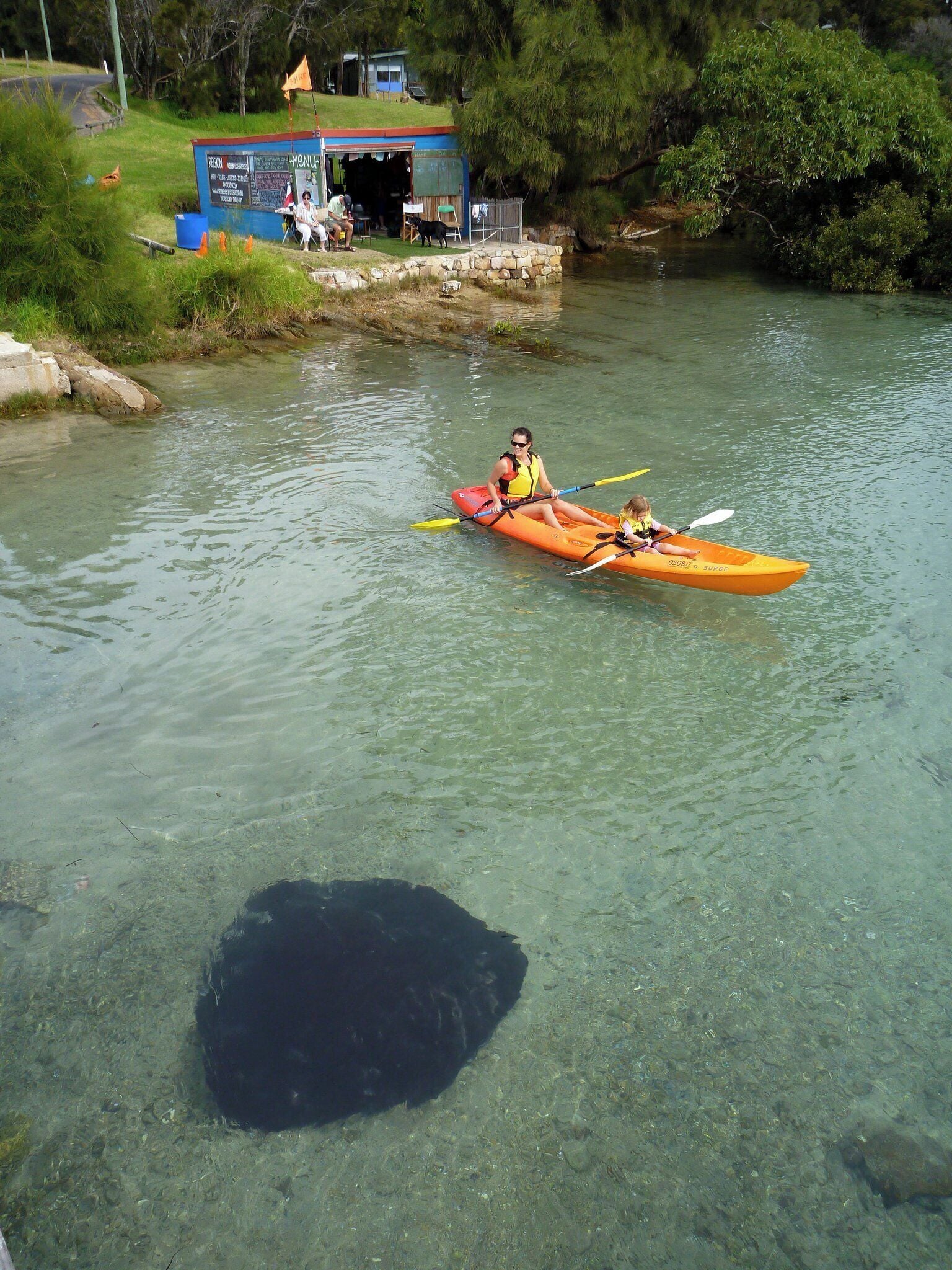 Un kayak sur une rivière, avec un quai et un petit bâtiment en arrière-plan.