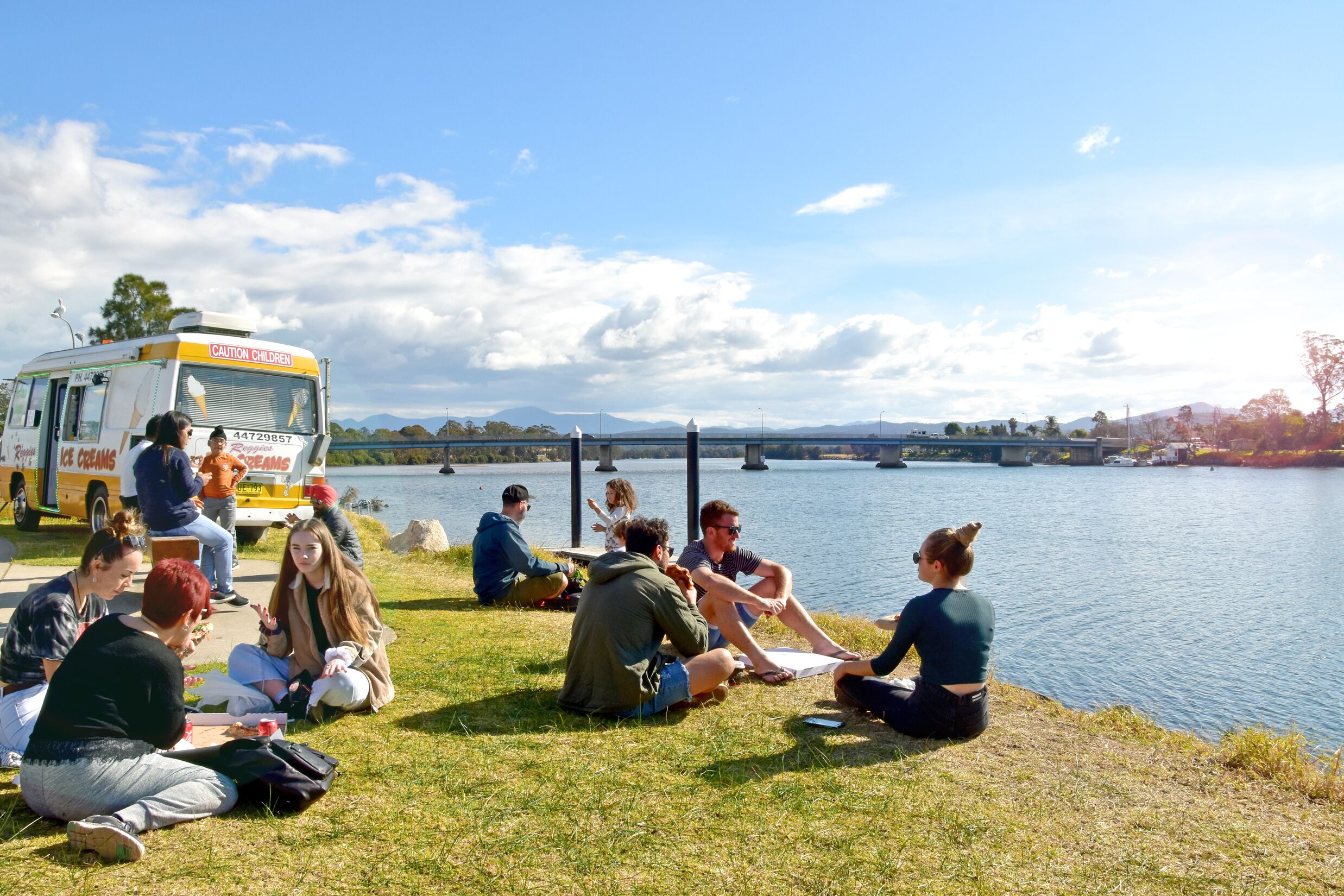 A group of people sitting on grass by a lake, with a food truck in the background.
