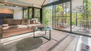 A modern living room with a sofa, coffee table, and large windows offering a view of the outdoors. - The Bower at Broulee (Broulee)