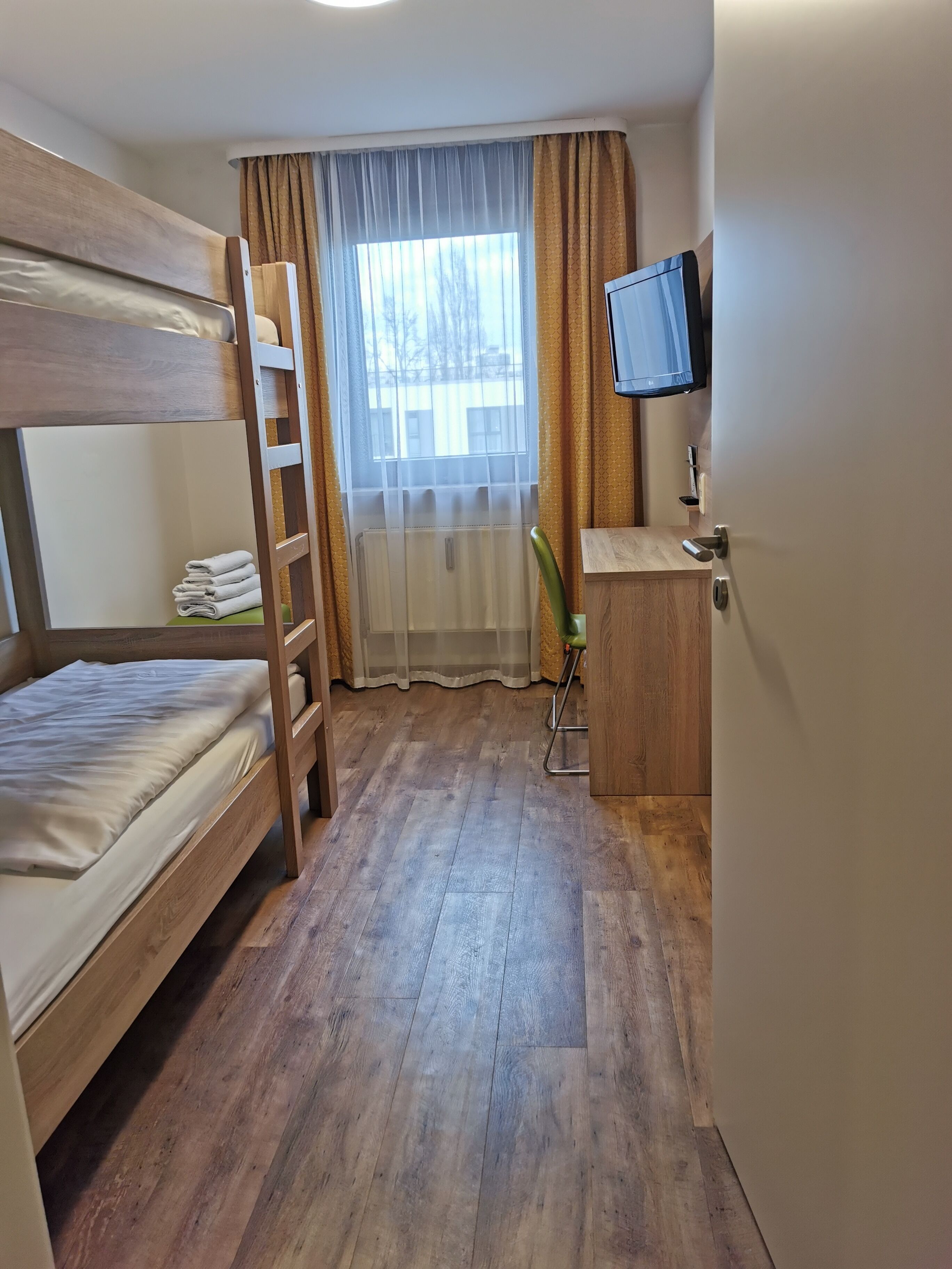 A bunk bed room with a wooden floor, a desk with a chair, a television, and a window with curtains.