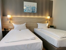 A hotel room with two beds, a wooden headboard, wall-mounted lamps, a painting, and a small plant.