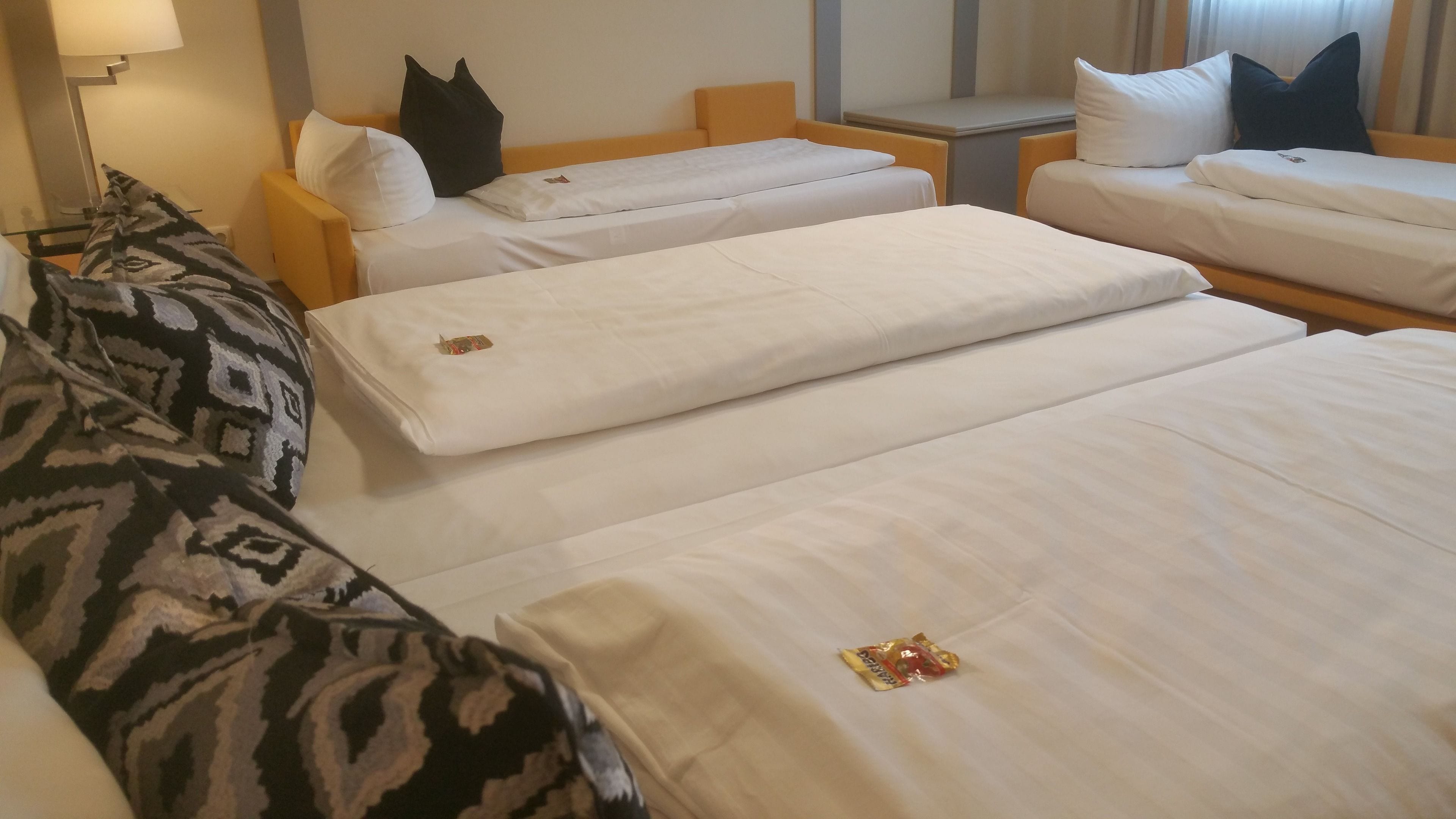 A hotel room with three beds, each with white linens and pillows. There is a small table with a lamp and a window with curtains.