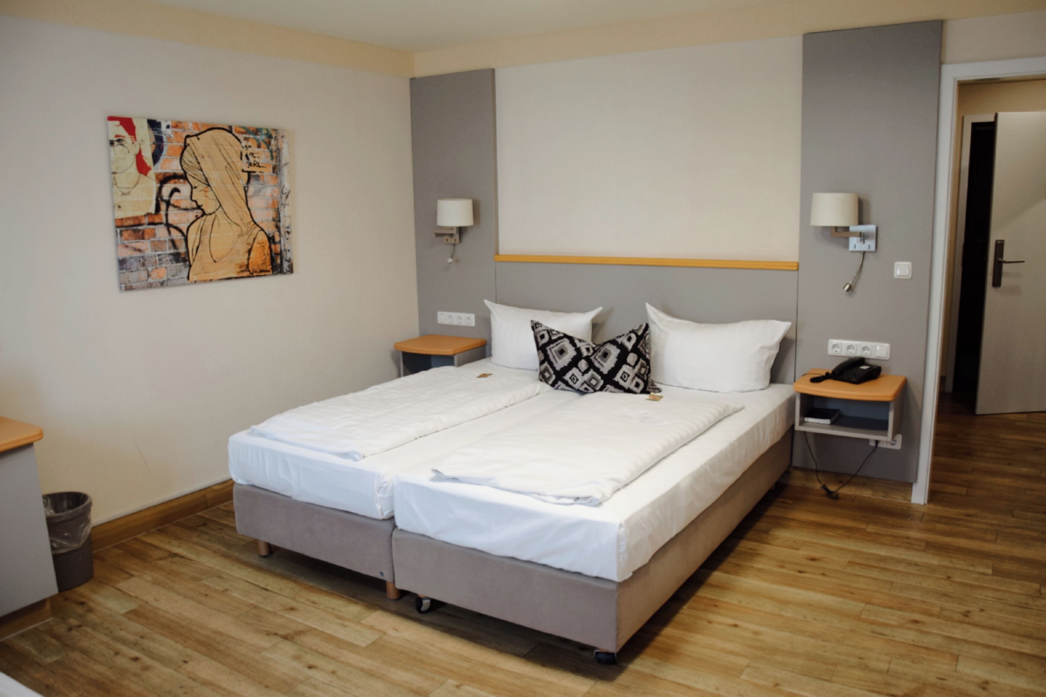 A double bed with a white and patterned headboard, two bedside tables with lamps, a small trash bin, and a painting on the wall.