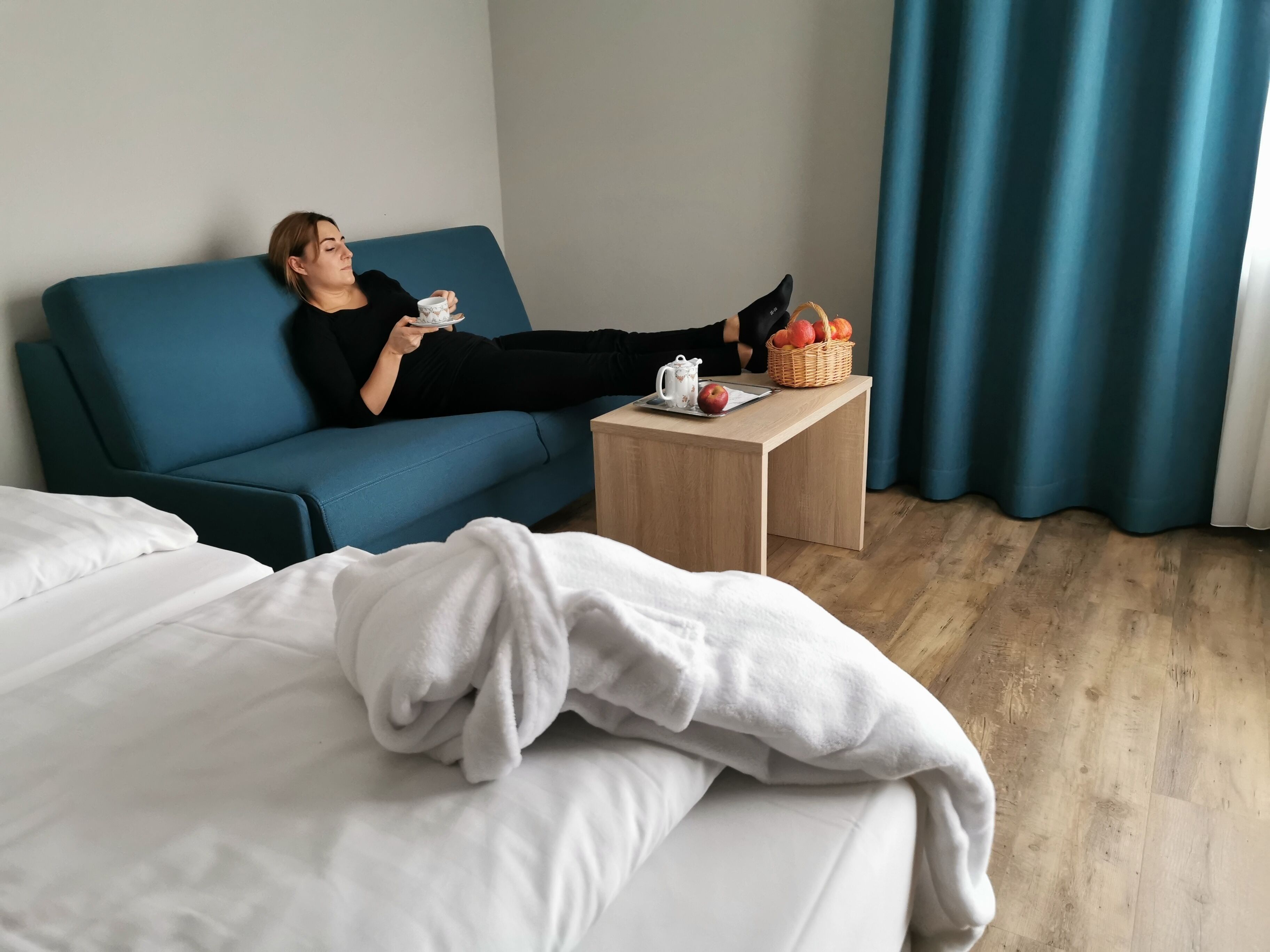 A person relaxing on a blue sofa with a basket of fruit and a kettle on a small table beside them.