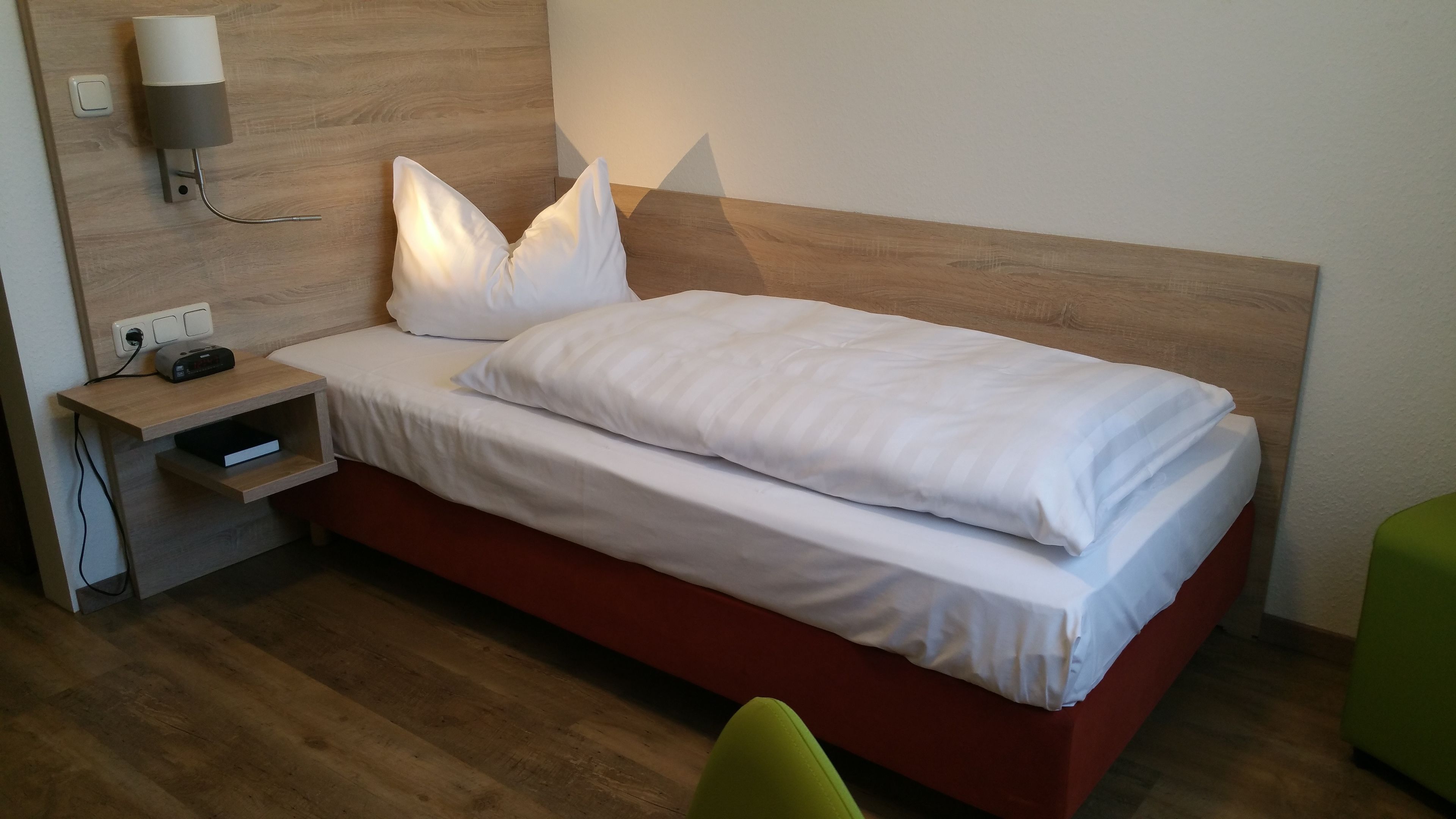 A neatly made bed with white linens, a bedside table with a lamp, and a power outlet.
