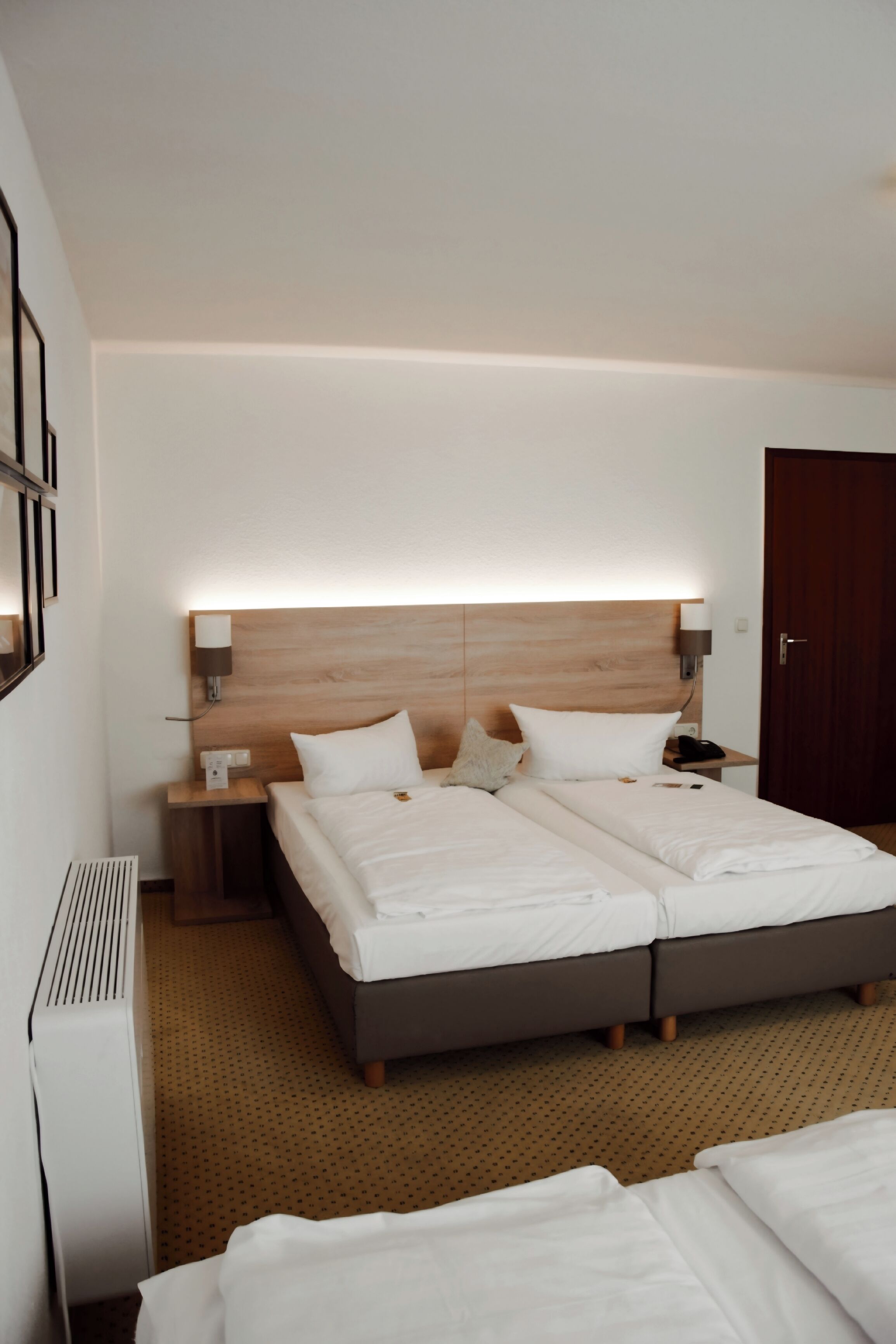 Habitación de hotel con una cama doble, una mesita de noche con lámpara, cabecera de madera y una puerta.
