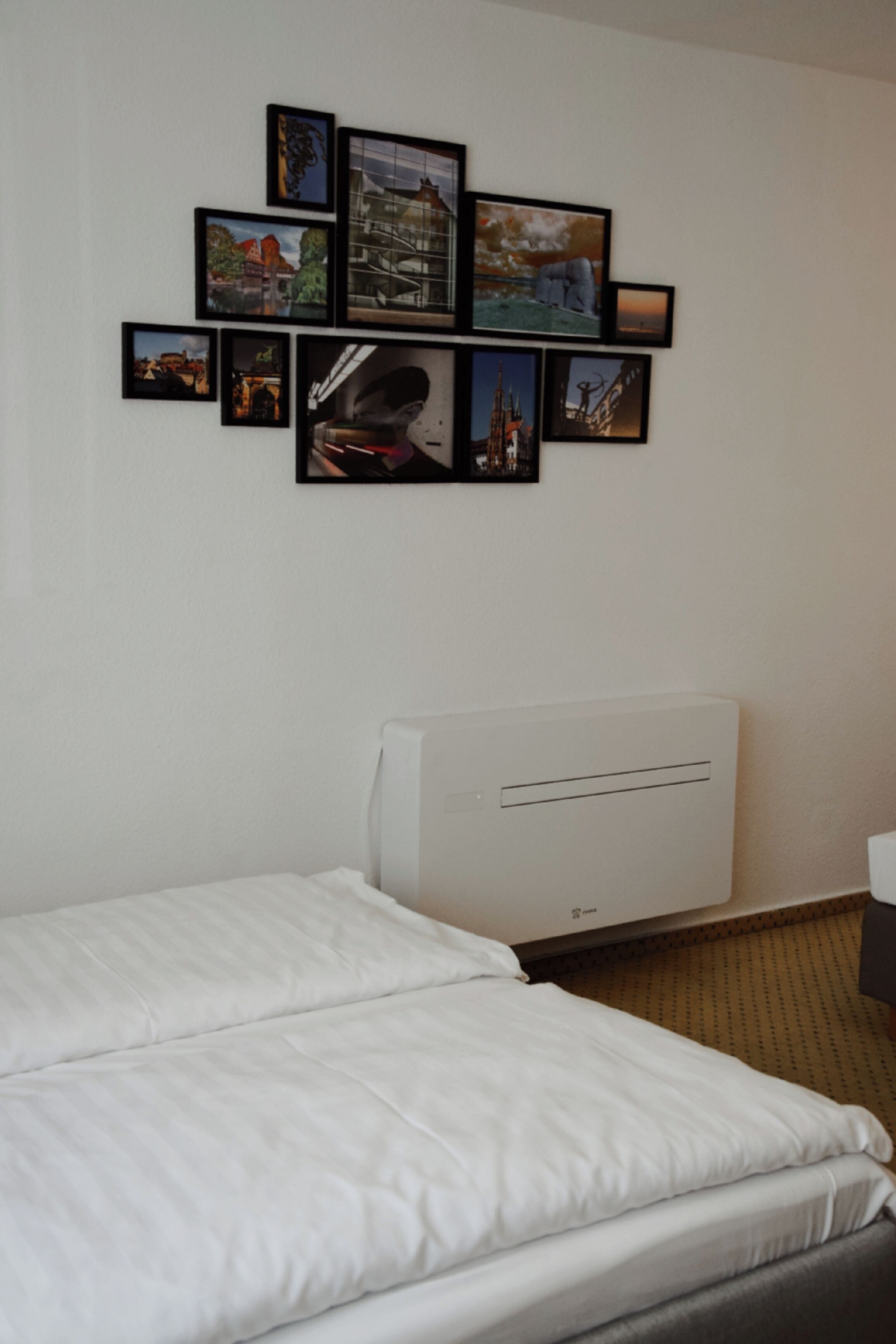 Habitación de hotel con cama, aire acondicionado de pared y varias fotografías enmarcadas.