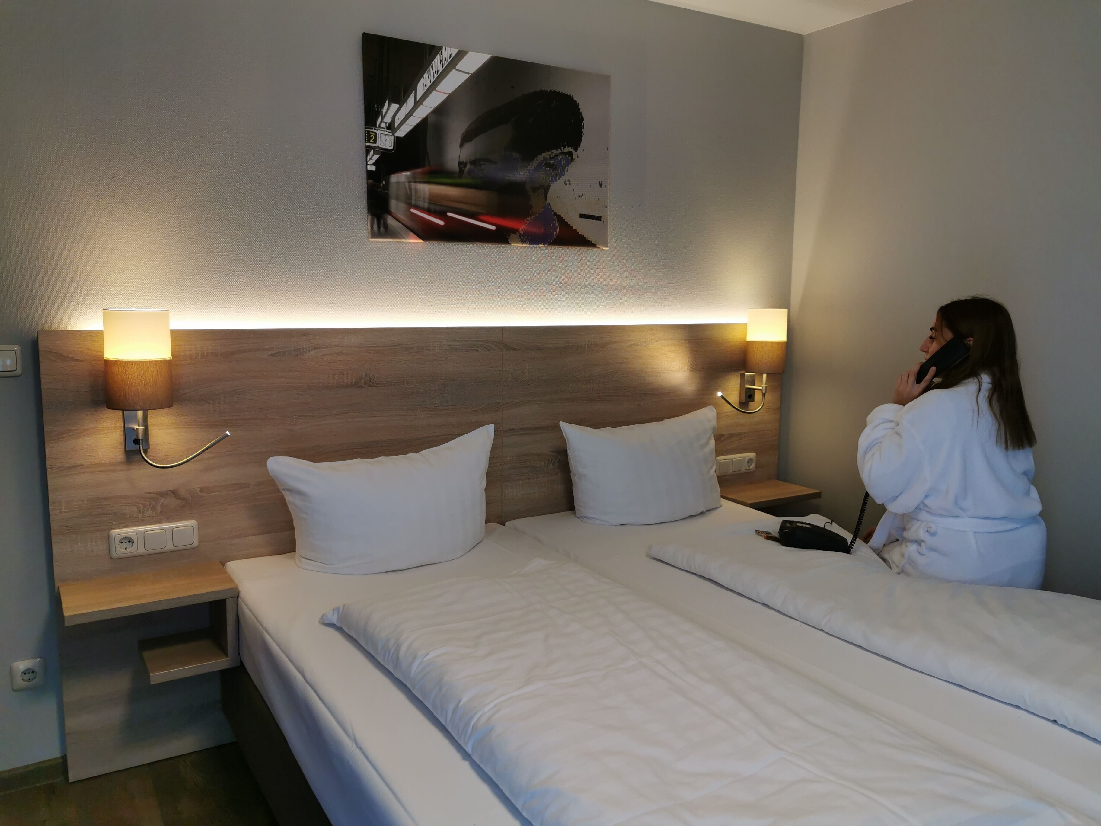 A hotel room with a wooden headboard, two bedside tables with lamps, a bed with white linens, and a painting above the bed.