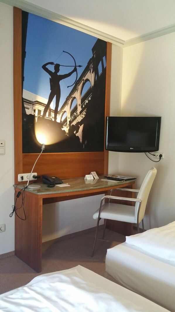 A hotel room with a large bed, a desk with a chair, a flat-screen TV mounted on the wall, and a painting of a bridge.