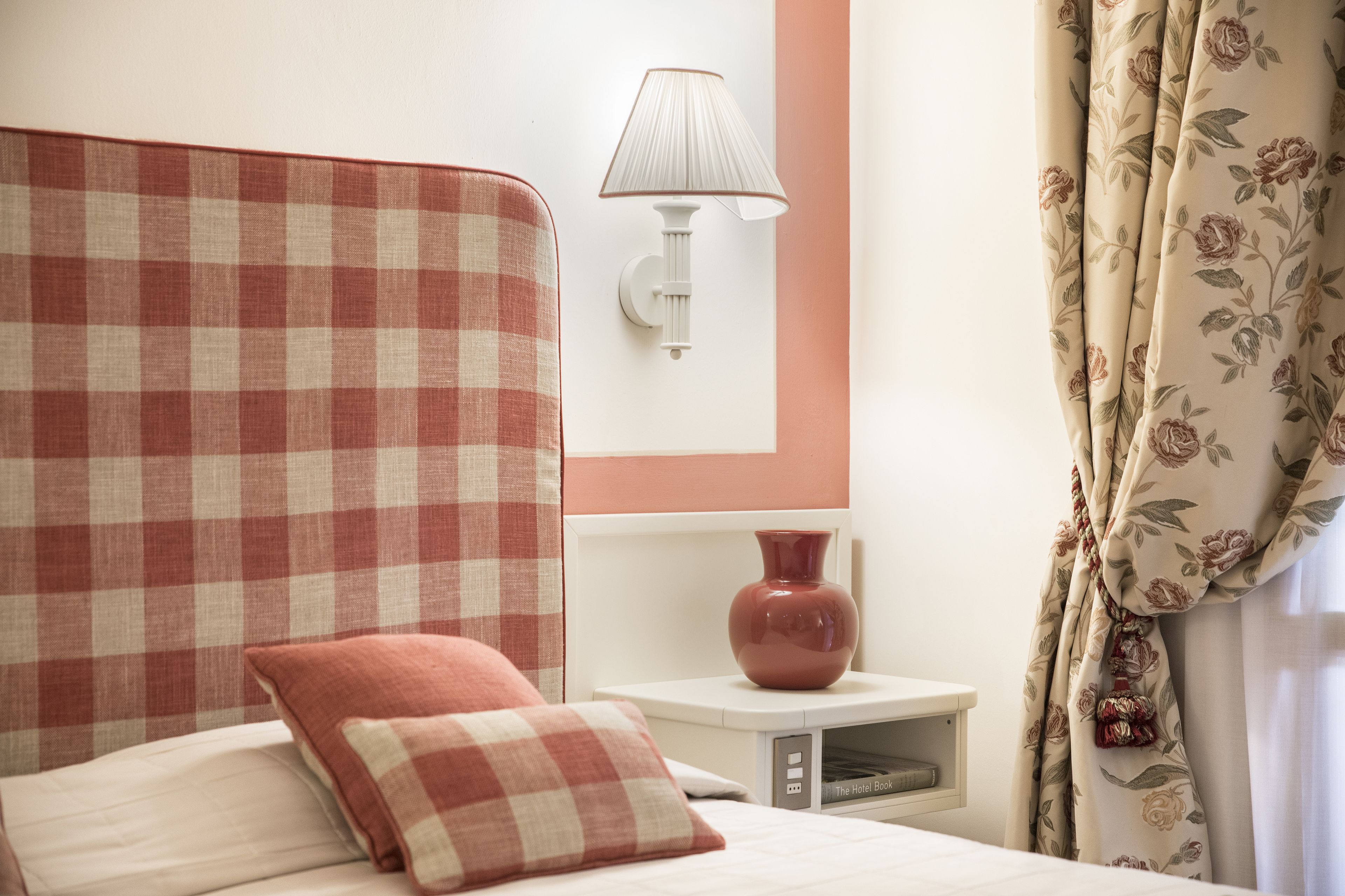 Habitación de hotel con una cabecera estampada, una lámpara de noche, un florero en la mesita y cortinas con motivos florales.