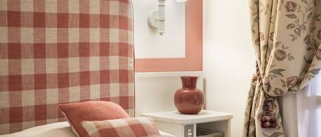 A hotel room with a checkered headboard, a bedside lamp, a vase on the nightstand, and floral curtains.
