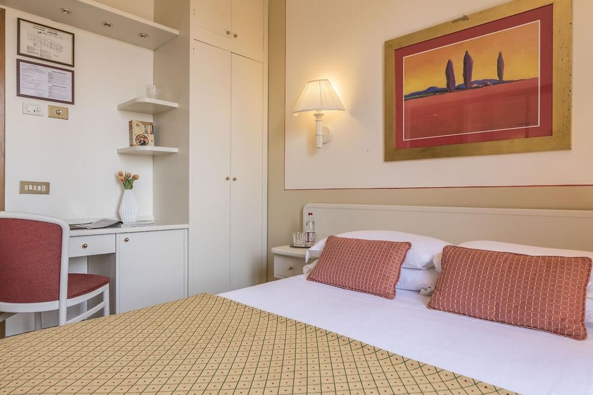 A hotel room with a bed, desk, chair, and a painting on the wall.