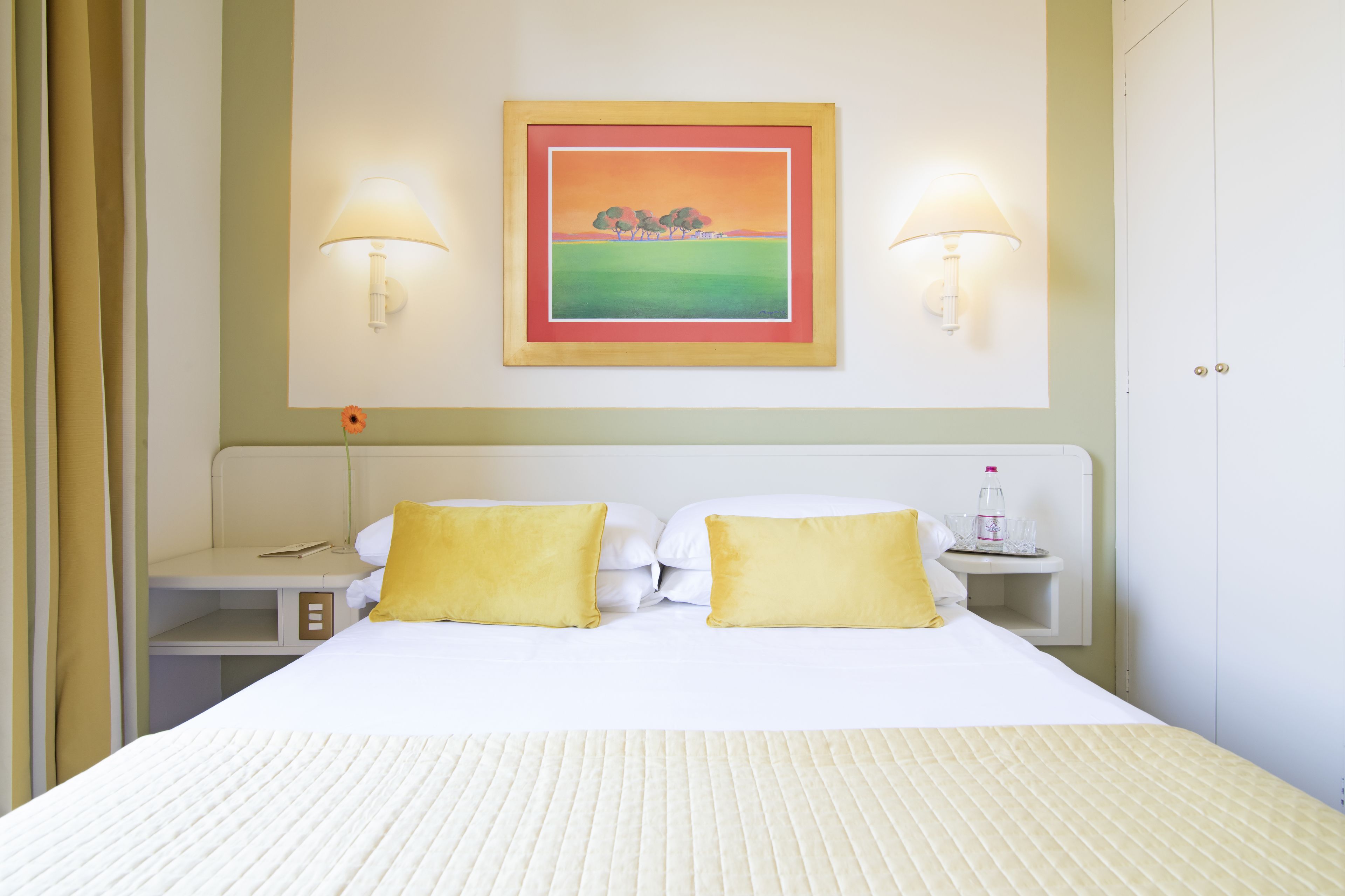 A hotel room with a bed, two bedside lamps, a nightstand, a painting on the wall, and a door.
