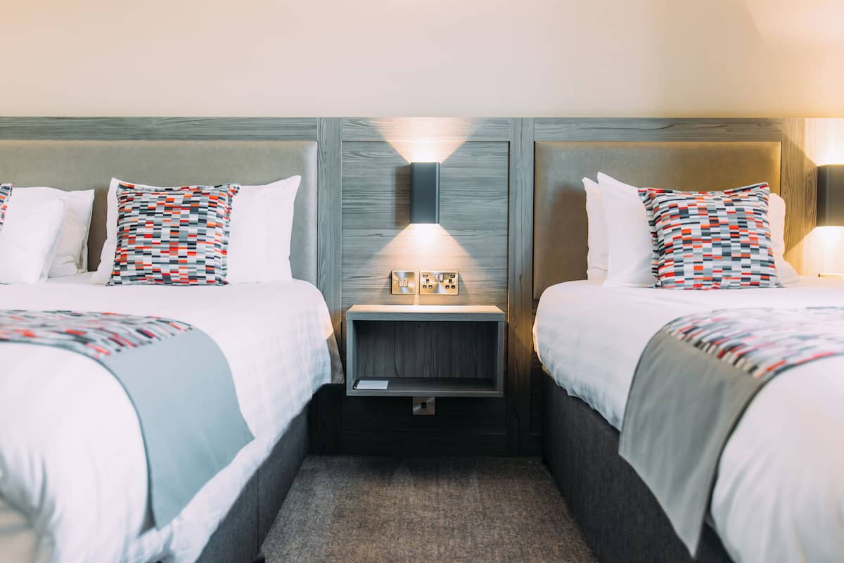 A hotel room with two beds, each with a patterned pillow, a bedside table with a lamp, and a wall-mounted light fixture.