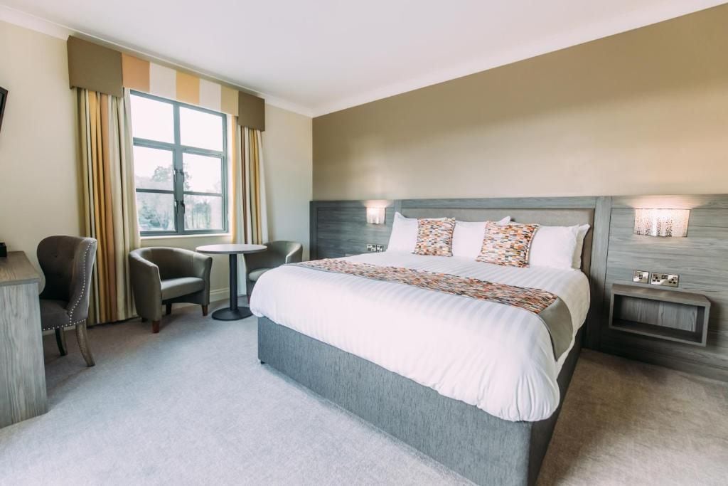 A hotel room with a large bed, a bedside table, a chair, a small round table, and a window with curtains.