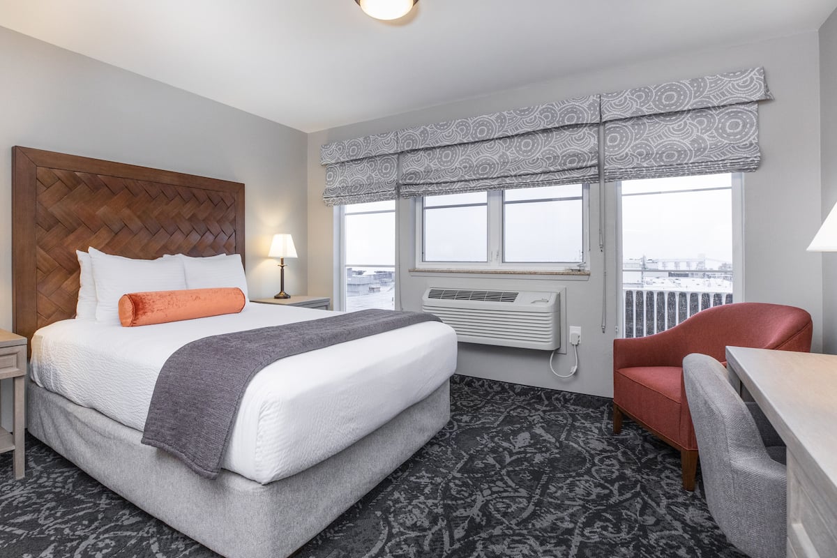 A hotel room with a large bed, a red chair, a desk, and a window with a patterned valance.
