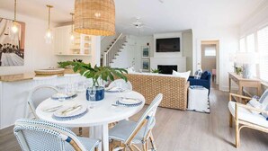 A modern kitchen with white cabinets, a dining area with a round table and wicker chairs, and a living area with a blue sofa and a fireplace. - The Wauwinet (Nantucket)