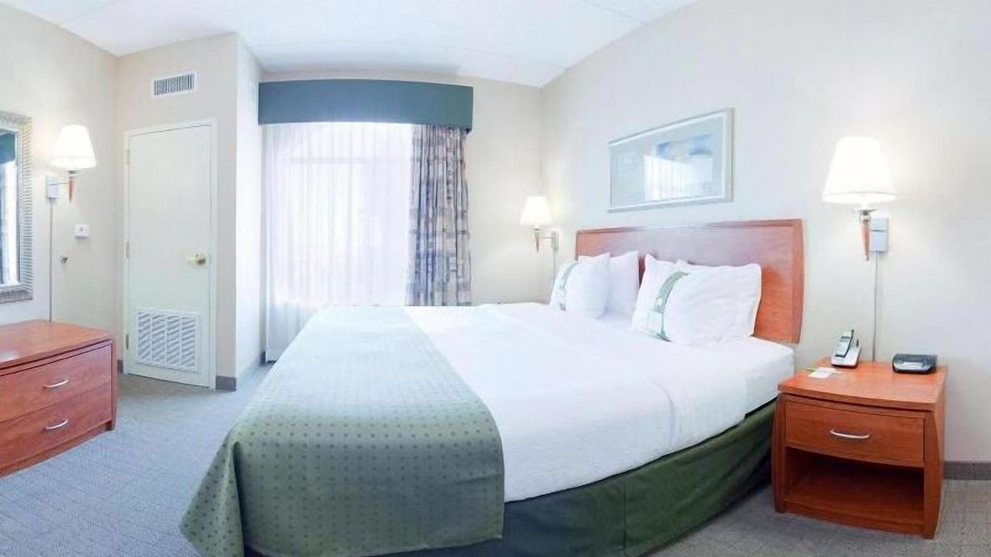 Una habitación de hotel con una cama, dos mesitas de noche, una lámpara de pared y una ventana con cortinas, además de un cuadro enmarcado.
