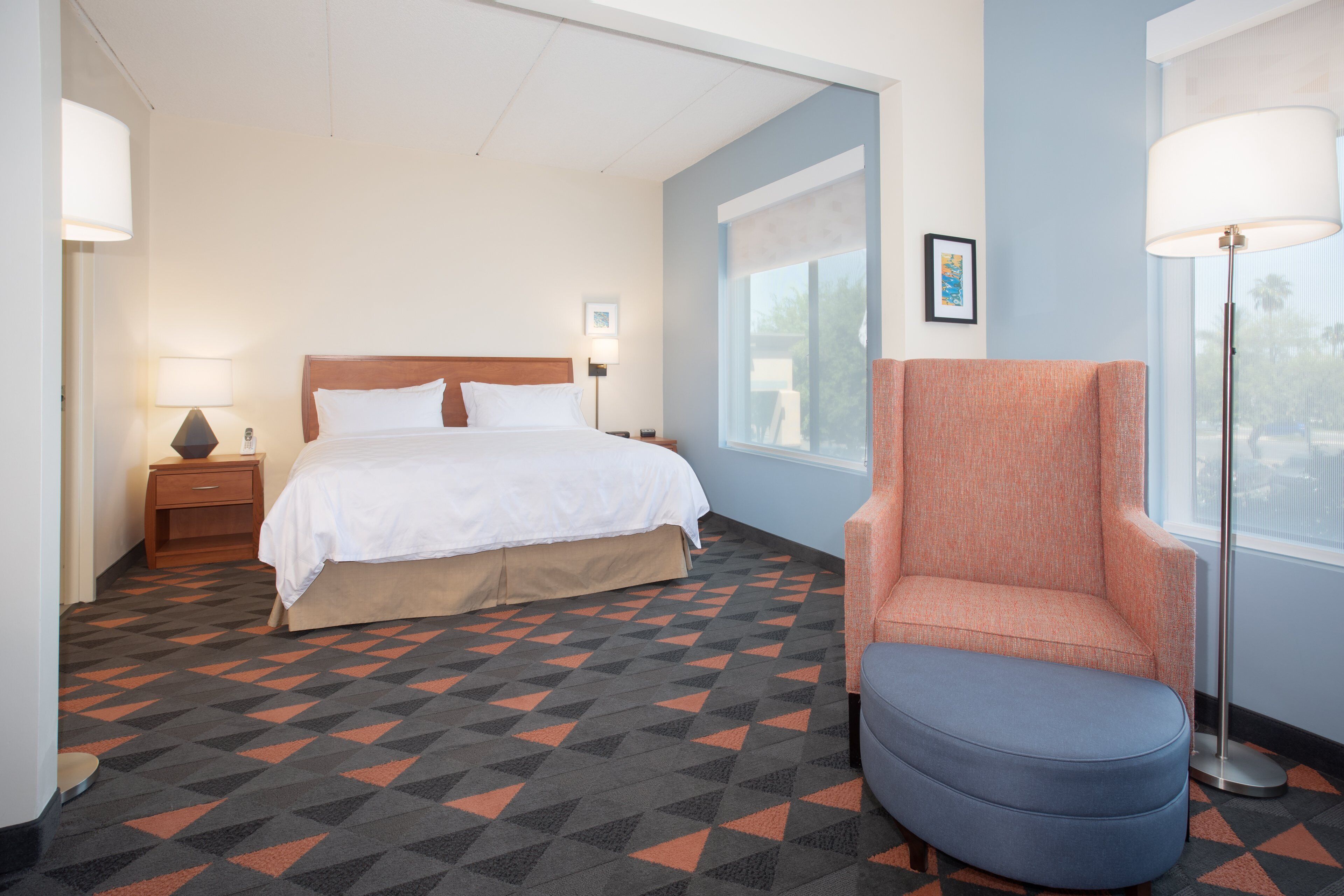 A hotel room with a bed, a chair, a nightstand, a lamp, and a window.