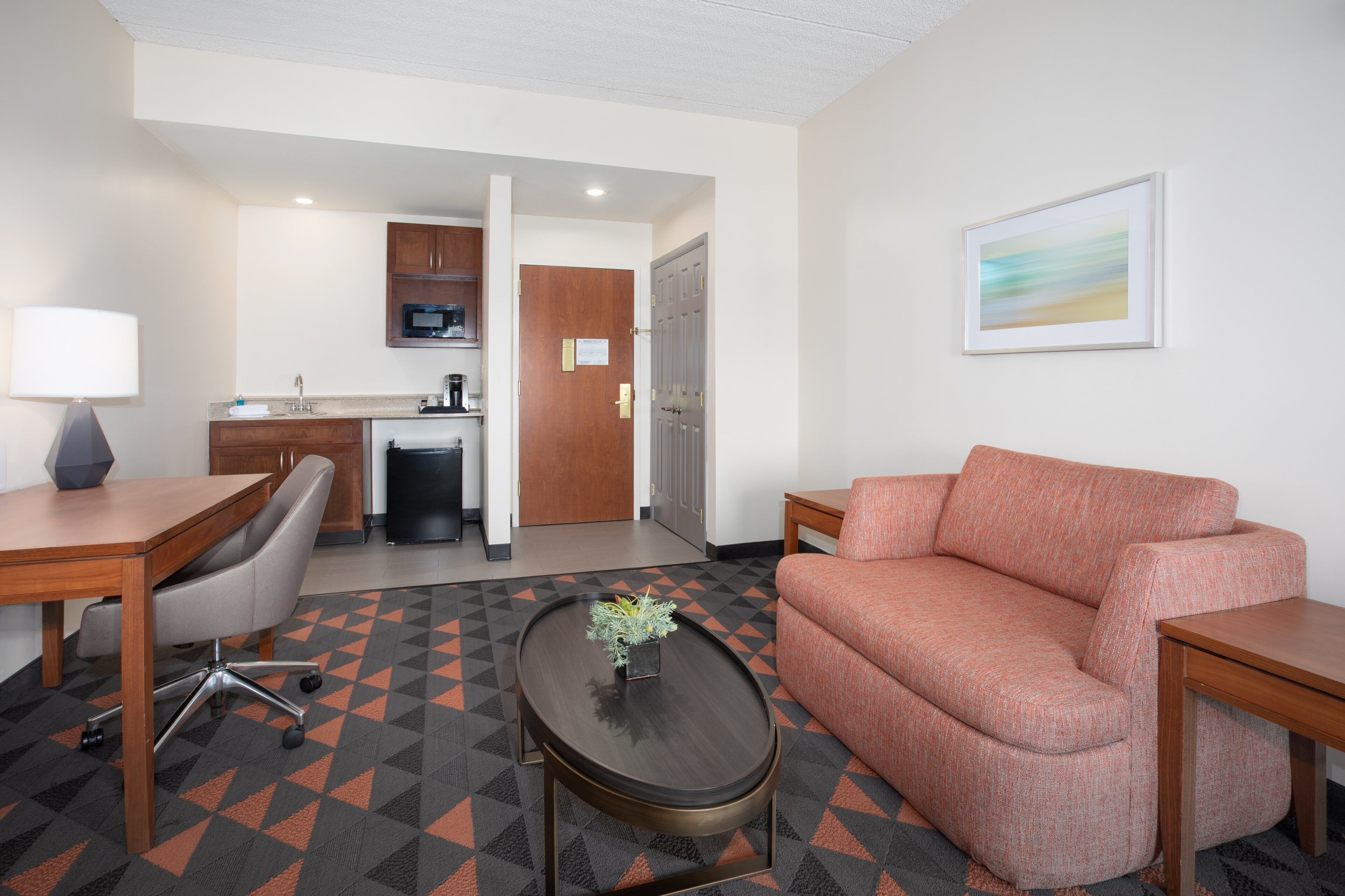 A hotel room with a sofa, a coffee table with a plant, a desk with a lamp, a kitchenette, and a door leading to another room.