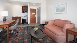 A hotel room with a sofa, a coffee table with a plant, a desk with a lamp, a kitchenette, and a door leading to another room. - Holiday Inn & Suites Goodyear - West Phoenix Area by IHG (Goodyear)