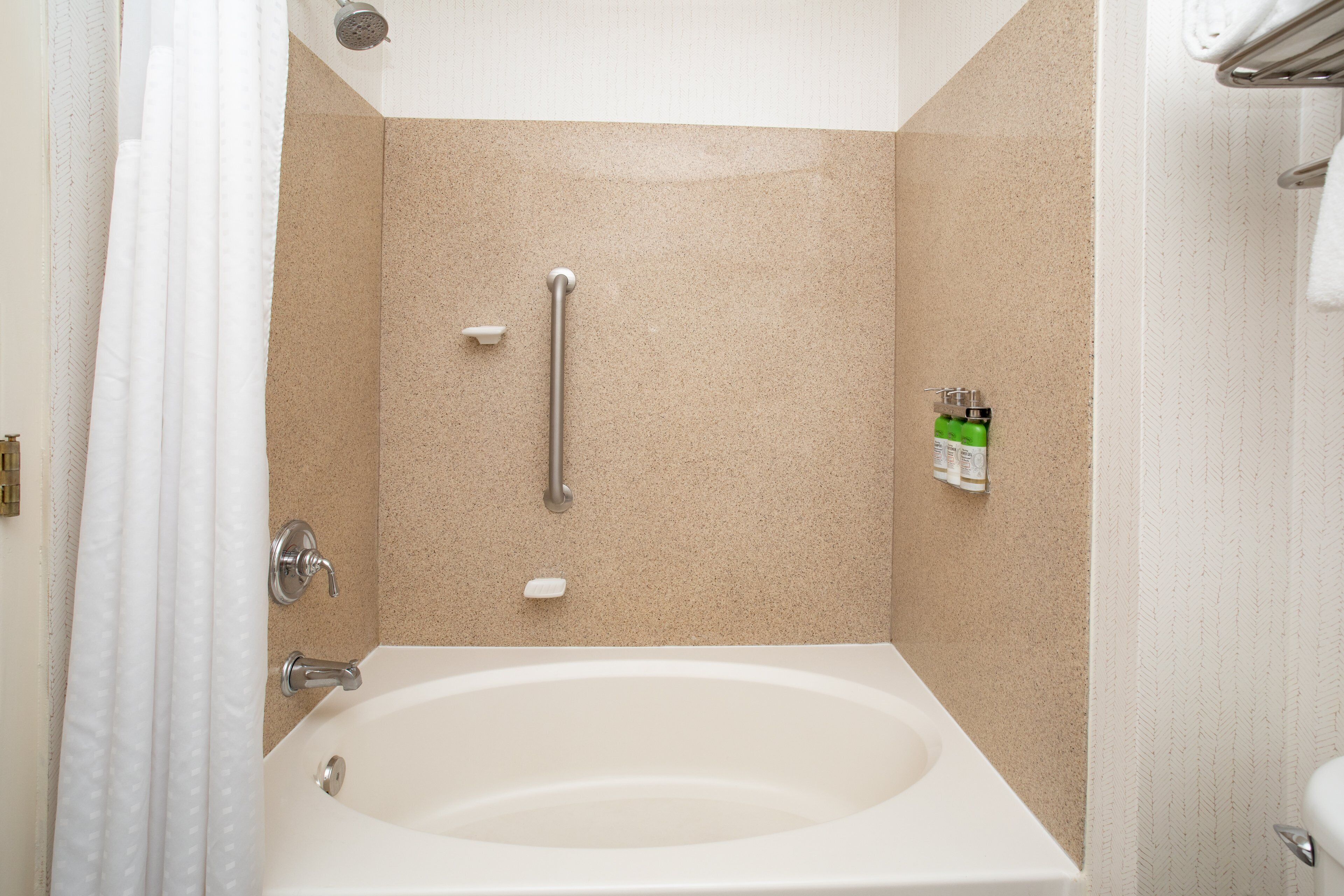 A bathroom with a bathtub, a handrail, and a soap dispenser on the wall.