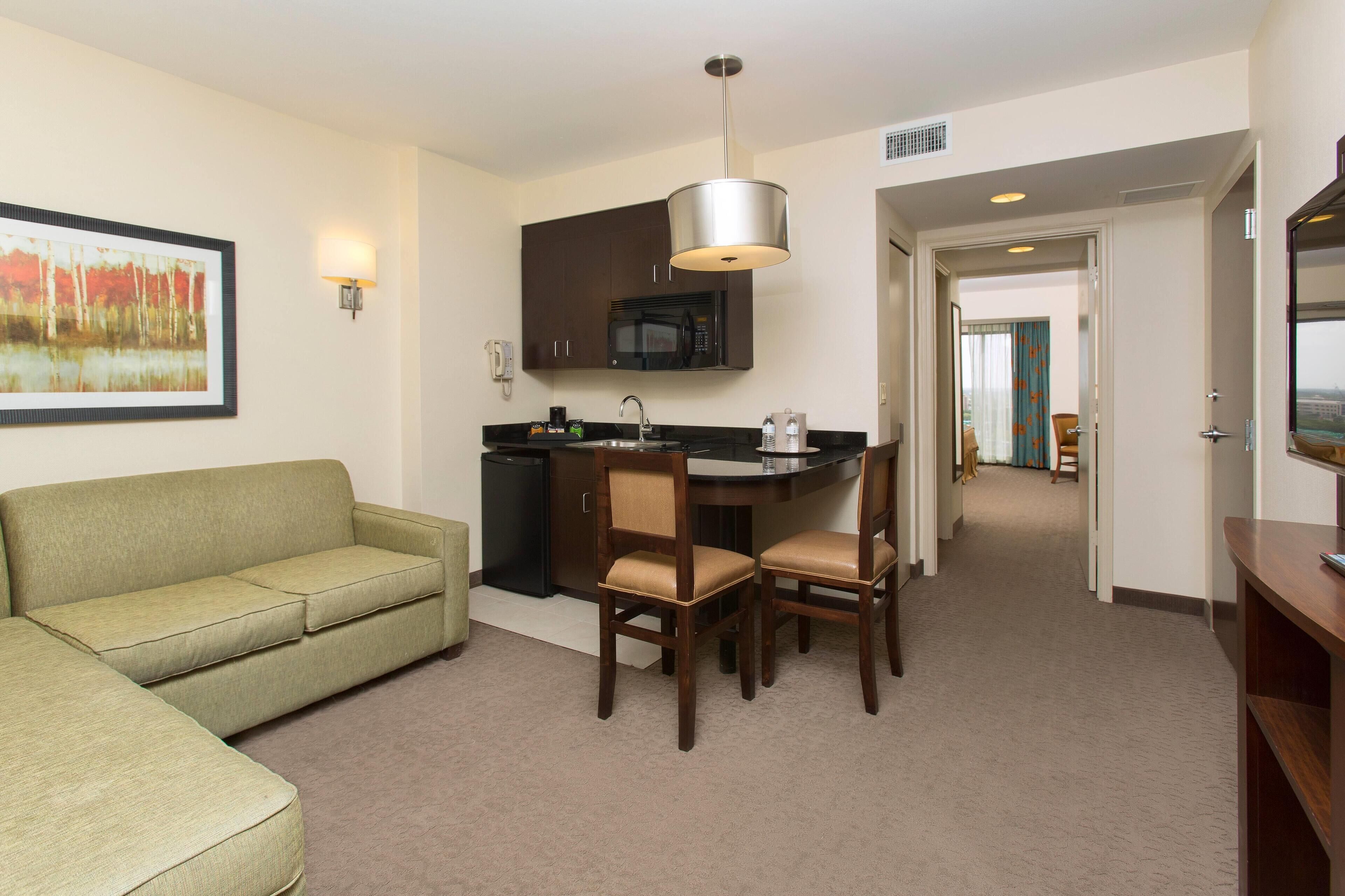A hotel room with a kitchenette, a dining area with a table and chairs, a sofa, and a painting on the wall.