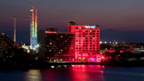 Ein Hotel mit mehreren Stockwerken, einem markanten Turm mit farbigen Lichtern und einem Gewässer im Vordergrund.