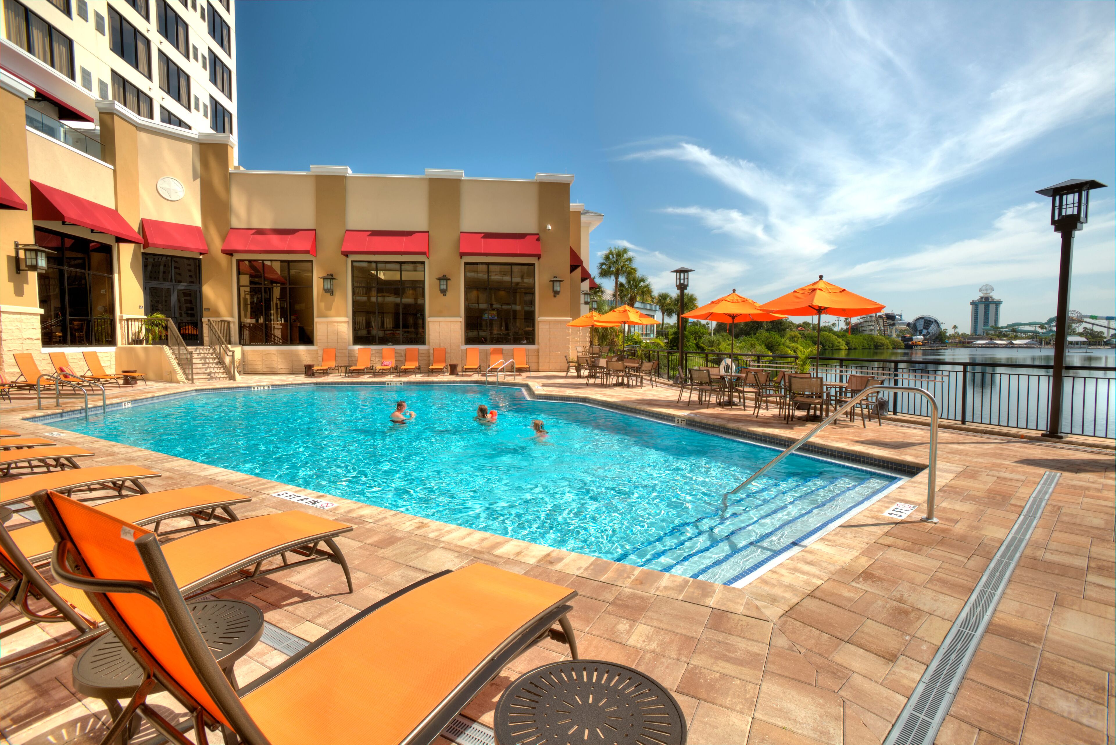 A swimming pool with lounge chairs and umbrellas, surrounded by a building with red awnings.