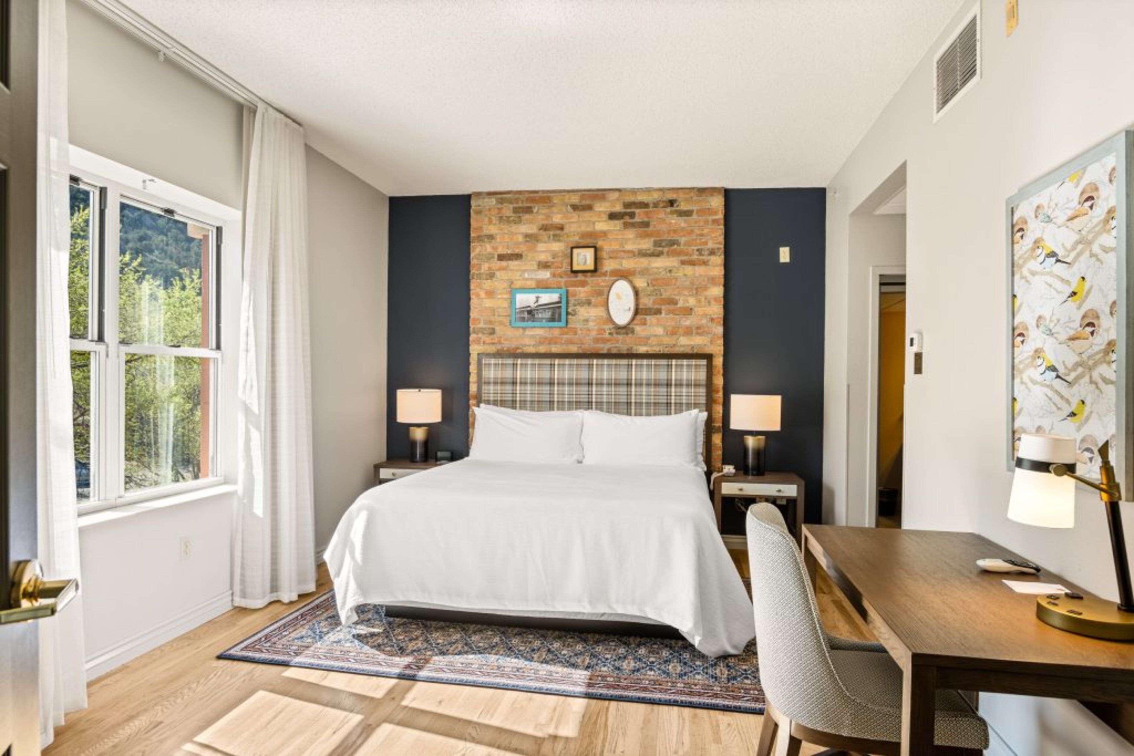 Une chambre d’hôtel avec un grand lit, un bureau en bois, une chaise, deux tables de chevet avec des lampes, un mur décoratif en briques et une fenêtre avec des rideaux.