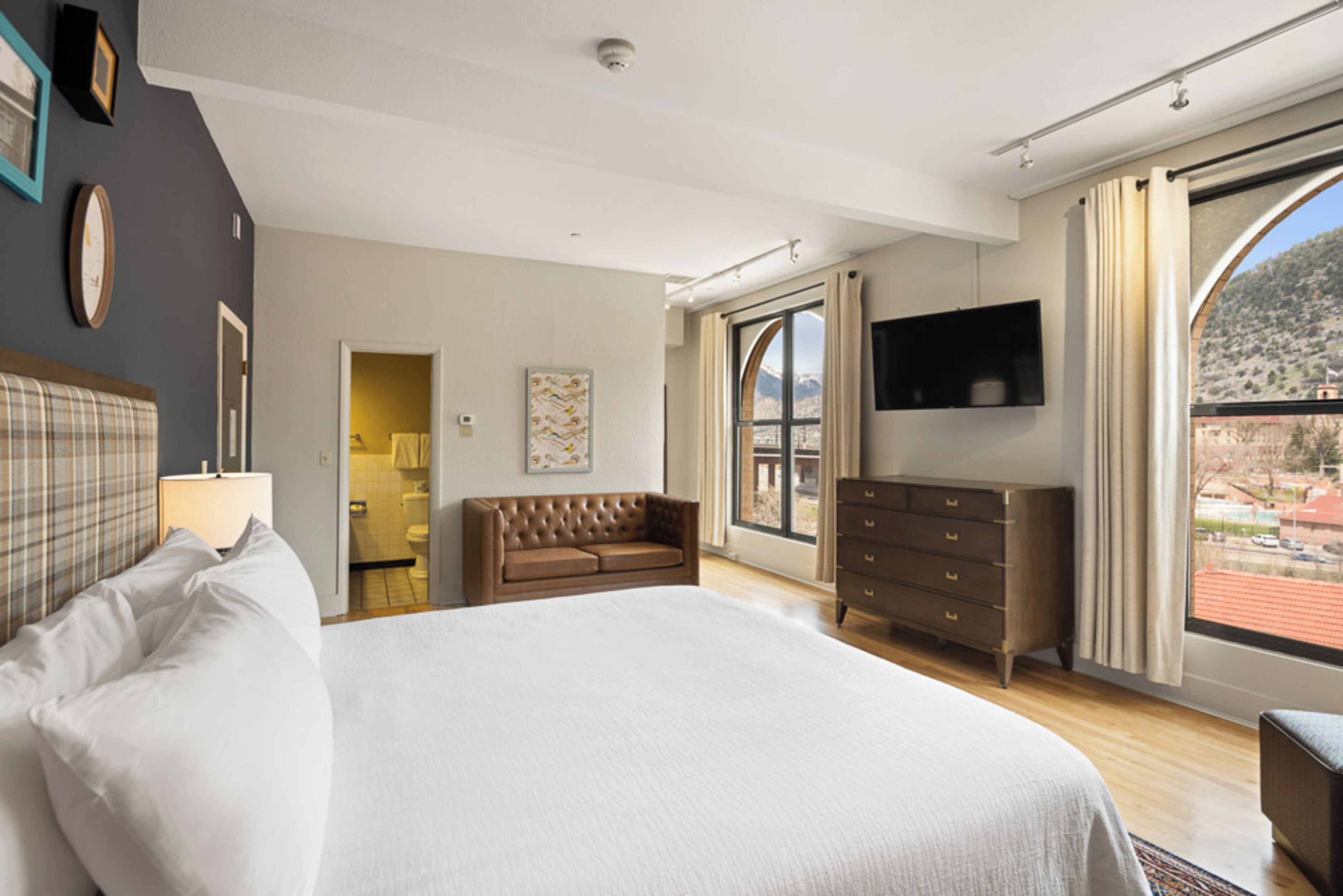 Quarto com uma cama grande, cômoda, televisão e vista para uma paisagem montanhosa através de uma janela ampla.