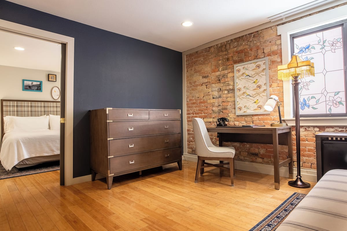 Un dormitorio con un aparador de madera, un escritorio con silla, una cama y una pared de ladrillo con obras de arte.