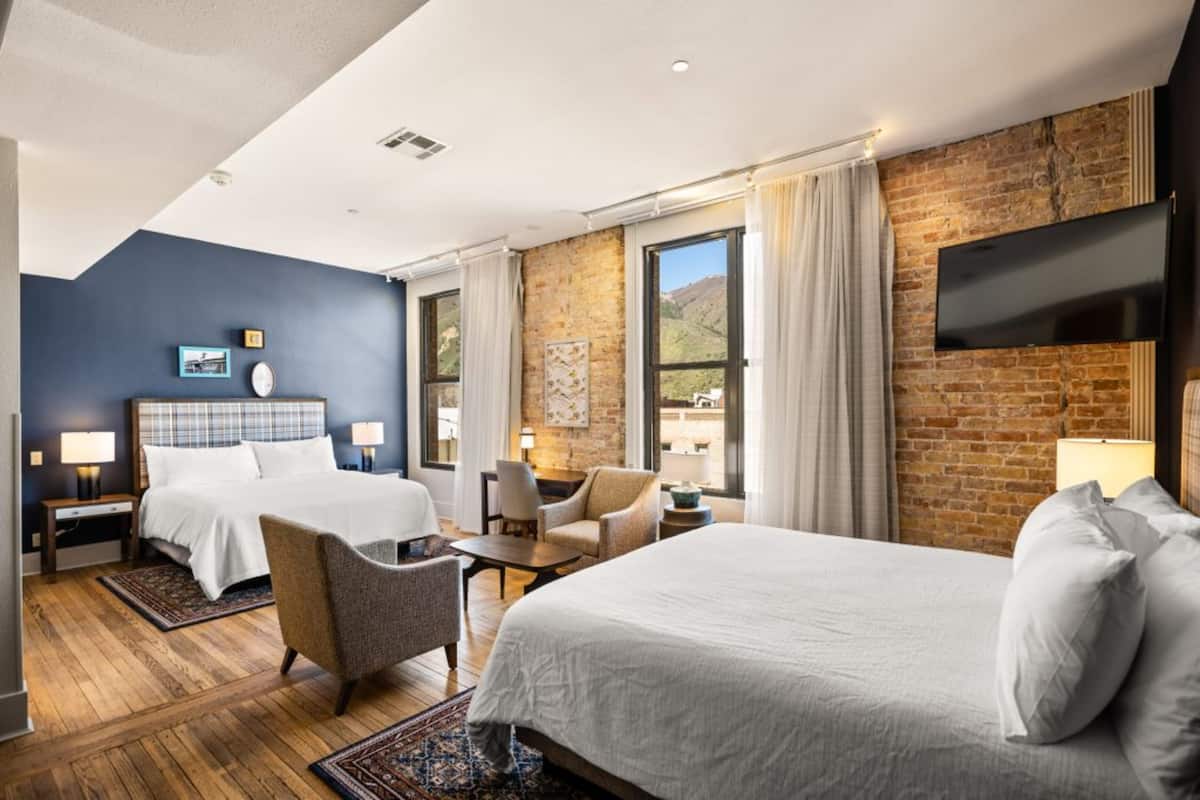 Habitación de hotel con dos camas, una pared de ladrillo, una televisión de pantalla plana y ventanas grandes.